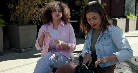 Two women chatting, searching purse at sunlit outdoor cafe with ar icons highlighting items