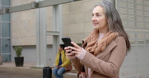 Mature woman standing with smartphone at transit stop wearing tan coat and knit scarf