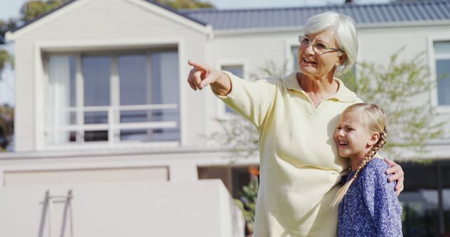 Senior woman hugging young granddaughter while pointing toward distance outside a house. Useful for concepts of family bond, intergenerational relationships, and enjoying quality time together. Can be used in advertisements and articles related to family life, senior living, retirement communities, and multigenerational families.