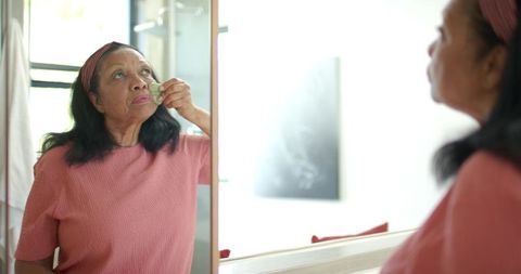 Senior african american woman applying gua sha facial massage in front of mirror