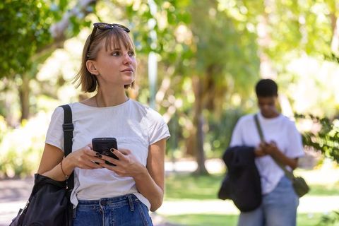 Young woman standing on sunlit park path holding smartphone with sunglasses and backpack