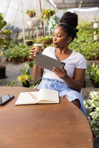 African american woman working on tablet in greenhouse with coffee and notebook