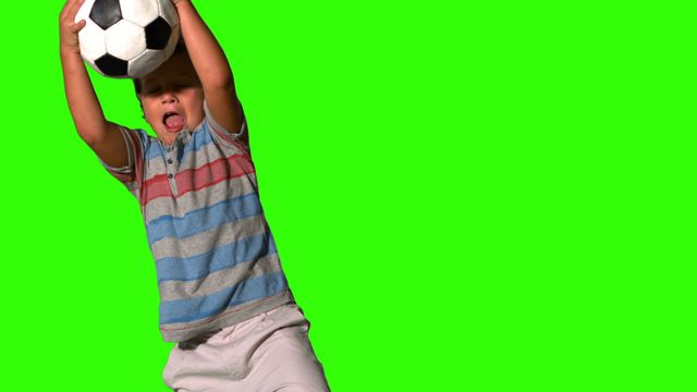 Little boy showing energetic pose while catching a soccer ball in front of a green screen. Great for sports-themed designs, children's activity promotions, or educational material emphasizing physical activity and fun.