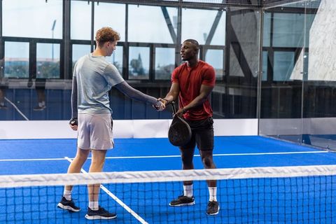 Diverse male padel players shaking hands on indoor blue court after match
