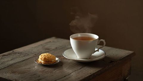 Steaming porcelain teacup with biscuit on weathered wooden table, rustic warm tea moment