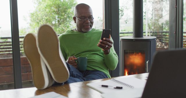 Man is sitting comfortably with his feet up, sipping coffee while using a smartphone. Windows and plants create a cozy, natural-lit environment. Ideal for themes of relaxation, digital lifestyle, home comfort, and remote work.