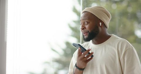 African man holding smartphone and using earbuds by window, casual home communication