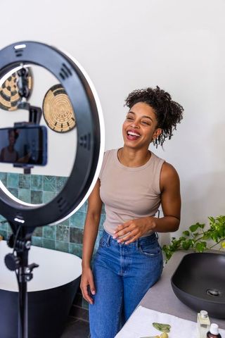 African american creator filming skincare routine by ring light at bathroom sink, smiling
