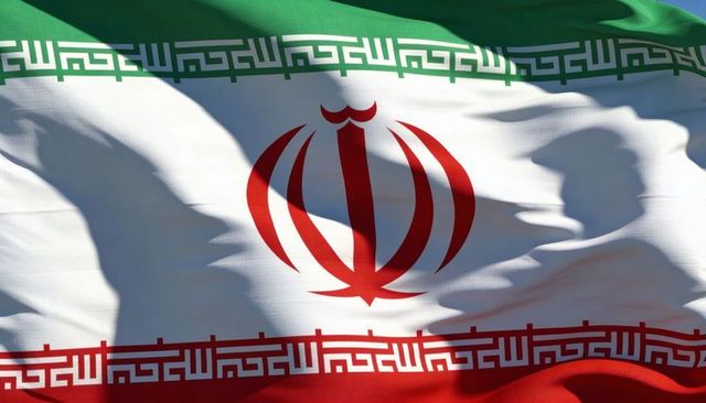 Waving iranian tricolor flag with central red emblem and kufic script bands, sunlit close-up