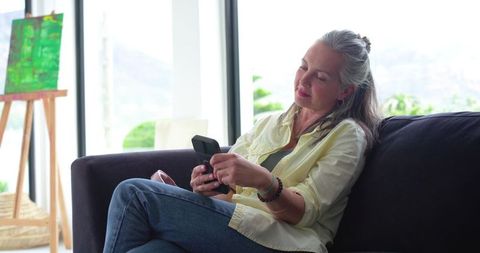 Reclining senior woman using flip phone on sofa in bright living room with easel