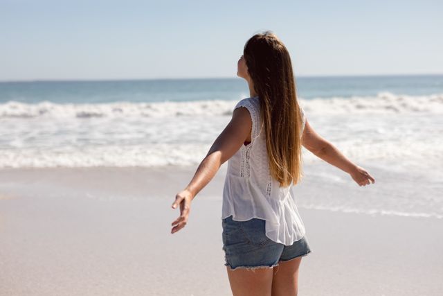 This image captures a woman standing on a beach with her arms outstretched, enjoying the sunshine and ocean view. Ideal for use in travel brochures, wellness blogs, lifestyle magazines, and advertisements promoting beach vacations, relaxation, and outdoor activities.