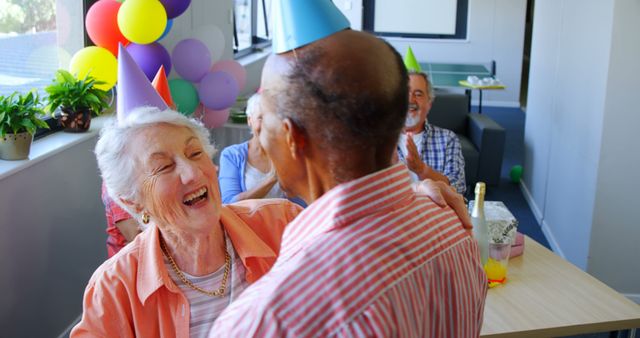 Senior adults are celebrating a birthday party, sharing joyful hugs. Vibrant balloons and party hats add a festive touch to the atmosphere. The happy expressions and warm interactions illustrate the essence of togetherness and friendship among elderly friends. Perfect for themes related to celebrations, elderly well-being, happiness, and social gatherings.