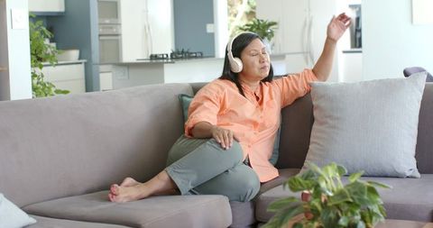 Mature woman relaxing on gray sectional sofa wearing headphones enjoying music at home