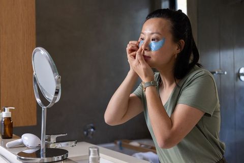Mature asian woman applying blue under-eye gel patches at bathroom sink mirror vanity