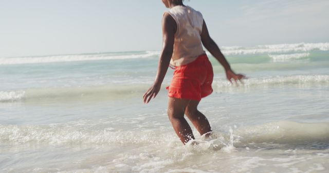 Child enjoying time at the beach splashing in shallow ocean water. Ideal for use in advertisements promoting family vacations, travel destinations, or summer activities. Can also be used in articles or blog posts about outdoor fun, childhood experiences, and beach days.