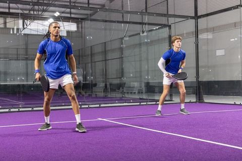 Diverse male padel teammates preparing to return serve on purple indoor court