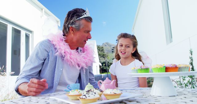 A middle-aged Caucasian man and a young girl are enjoying a playful tea party outdoors, with the man humorously dressed in a tiara and feather boa. Their joyful interaction over cupcakes creates a heartwarming scene of family bonding.
