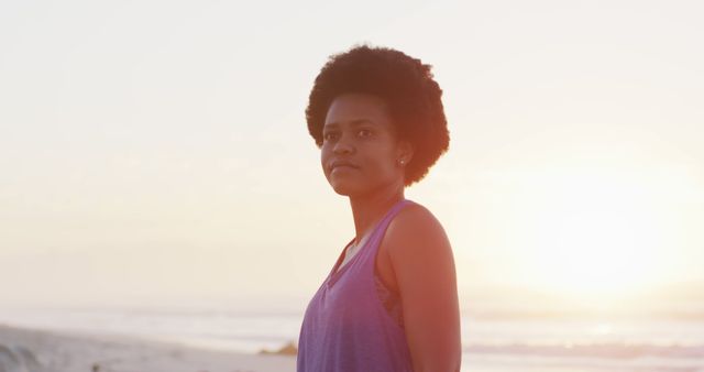 Young woman with afro hairstyle stands on the beach at sunset, radiating confidence and tranquility. Ideal for use in advertising related to self-confidence, beauty, natural landscapes, leisure activities, or wellness. Perfect for inspirational articles, blog posts, and social media content about nature, empowerment, and self-care.