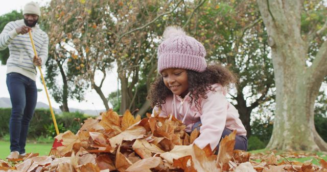 Young girl joyfully playing in a pile of autumn leaves with her father in the background raking. Perfect for illustrating family fun, seasonal activities, or advertisements focused on family time and outdoor enjoyment. Can be used in marketing campaigns for outdoor apparel, family-oriented events, or autumn-themed promotions.