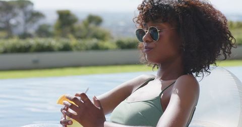 Relaxing african woman reclining on pool float wearing sunglasses sipping citrus drink