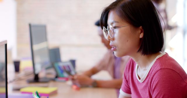 Young professional woman with glasses concentrating on monitor in modern office environment. Suitable for illustrating themes like tech industry, female professionals, workplace focus, and productivity.