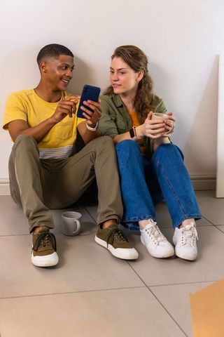 Diverse couple sitting on tiled floor sharing smartphone and coffee during casual move-in