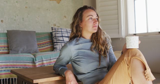 Caucasian woman sitting on floor in living room drinking coffee, looking out of window and smiling. simple living in an off the grid rural home.