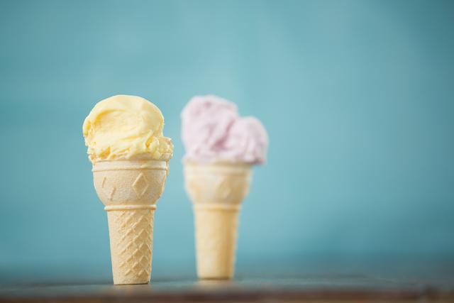 Two ice cream cones, one with vanilla and the other with strawberry flavor, standing on a wooden board against a blue background. Ideal for use in advertisements for ice cream shops, summer promotions, dessert recipes, and food blogs. The vibrant colors and simplicity make it perfect for social media posts and marketing materials.