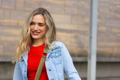 Smiling woman wearing denim jacket and red top with olive crossbody strap on urban street