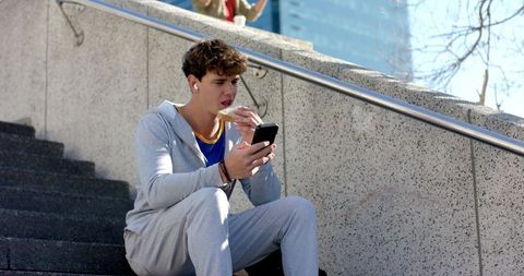 Young man eating sandwich while checking smartphone on urban steps with earbuds