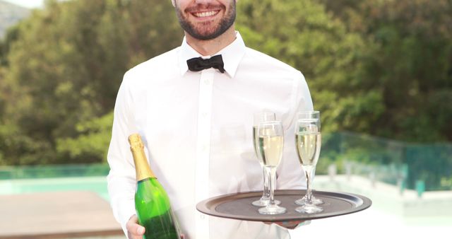 A smiling Caucasian waiter in a formal white shirt and bow tie holds a tray with champagne glasses and a bottle, with copy space. His professional attire and the outdoor setting suggest a celebratory event or upscale service.