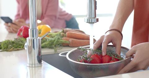 Couple rinsing tomatoes in colander under stainless faucet with fresh produce