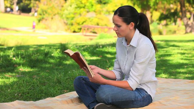 A young woman is reading a book while sitting on a picnic rug in a park. She appears relaxed and calm, enjoying the natural surroundings. This could be used for concepts of leisure, relaxation, education in nature, or enjoying outdoor activities. It is perfect for lifestyle blogs, articles, or advertising focused on reading, relaxation, or outdoor activities.