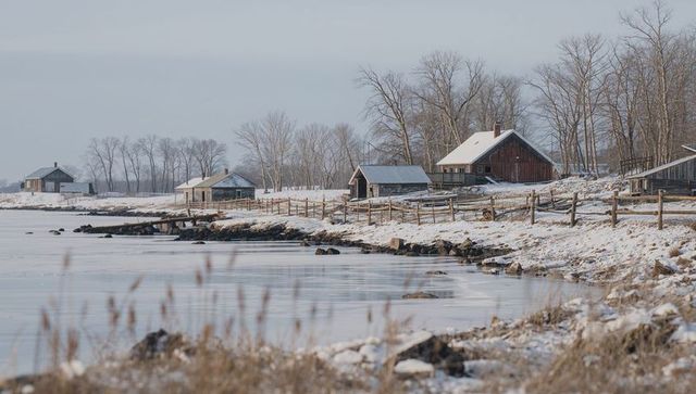 Lakeside farm buildings sitting behind split-rail fence on snowy frozen shoreline
