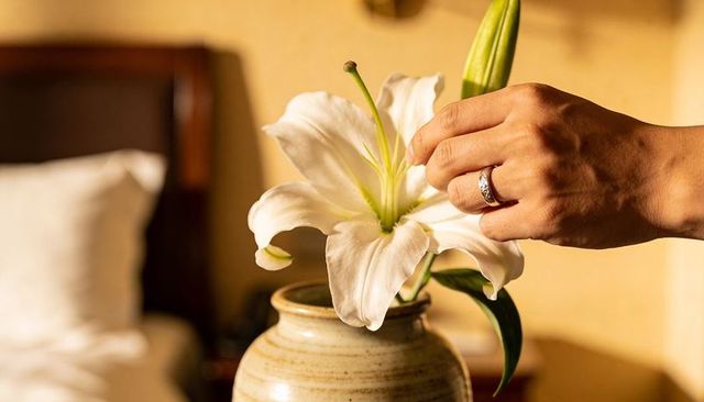 Male hand arranging white lily in beige ceramic vase on bedside table with silver ring