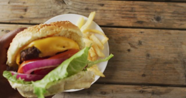 Cheeseburger with fresh lettuce, tomato, cheese, and onion in sesame bun held in hand. Plate of fries in background on rustic wooden surface. Perfect for use in articles or advertisements about food, recipes, fast food chains, or comfort foods.