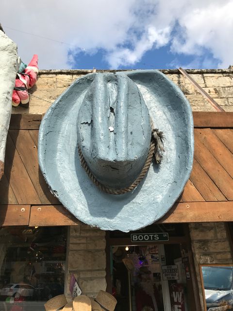 This image shows a large cowboy hat sculpture mounted above a rustic western-themed storefront. The weathered hat contrasts slightly with the blue sky and brick building. Ideal for themes related to western culture, Texas, rural stores, tourism, and outdoor markets.