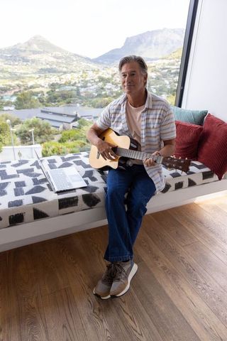 Mature man playing acoustic guitar on window seat with laptop overlooking sunlit valley