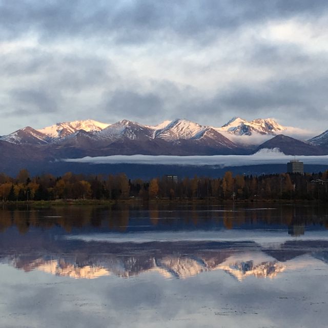 A panoramic view of snow-capped mountains reflected in a calm lake, surrounded by autumn foliage. This serene and majestic landscape is ideal for use in travel brochures, posters promoting nature-related activities, desktop wallpapers, and greeting cards.