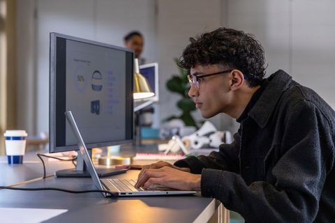 Focused asian man in 20s typing on laptop at modern coworking desk with monitor showing analytics