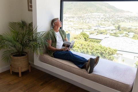 Mature man reclining on window bench reading and gazing out at sunlit suburban view