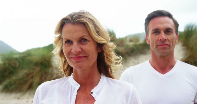 Mature couple smiling standing at the beach during a calm day. Man and woman dressed in casual white clothing enjoying the relaxed natural environment. Great for lifestyle, togetherness, outdoor activity, retirement, and health-related content with emphasis on happiness and bonding.