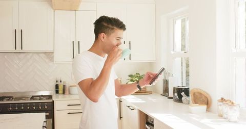 Asian man drinking from light green mug while checking smartphone in bright modern kitchen