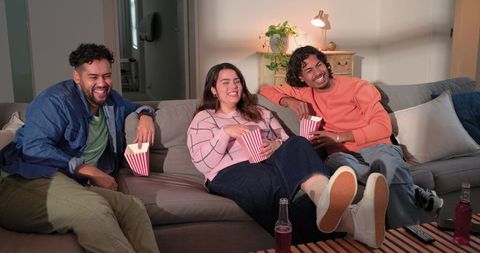 Three Friends Laughing and Snacking on Sofa During Cozy Movie Night at Home