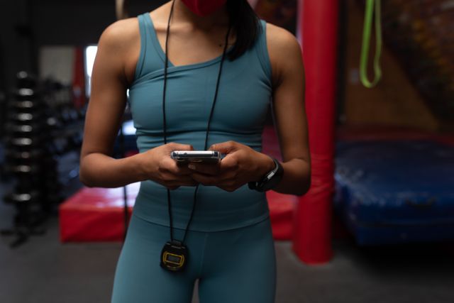 Caucasian woman in gym wearing a stopwatch around her neck and a facemask, typing on her phone. Ideal for use in fitness, health, and technology-related content, emphasizing active lifestyle, safety, and modern gym environments.