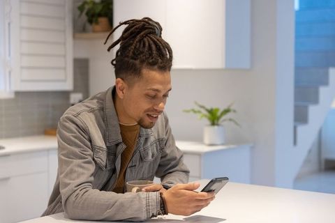 Mid-adult african american man checking smartphone at modern kitchen island holding mug