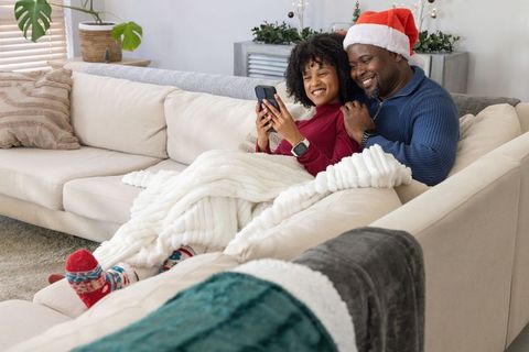 African american couple snuggling under blanket wearing santa hats and using phone on couch