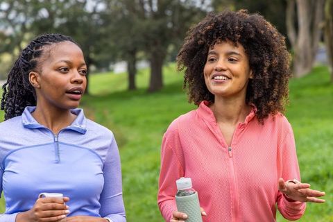 Diverse women walking and chatting in park holding reusable bottle and coffee cup