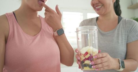 Diverse women preparing and tasting fruit smoothie in bright home kitchen