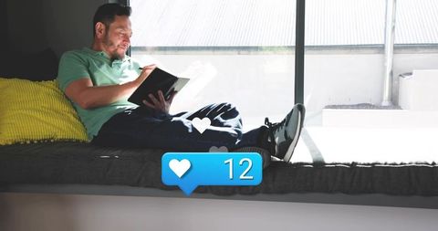 Reclining man on window seat using tablet with stylus and floating heart like notification